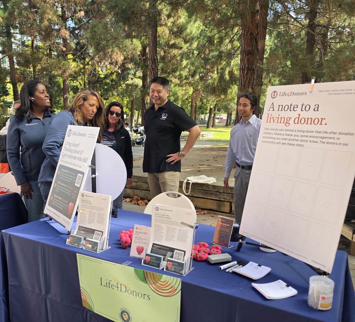 Visitors gather around the Life4Donors booth beneath trees, beside a spin wheel and a board inviting notes to living donors